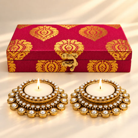 Gold-plated diya holders with sparkling crystals next to a red and gold patterned box for Diwali festival gifts