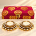 Gold-plated diya holders with sparkling crystals next to a red and gold patterned box for Diwali festival gifts