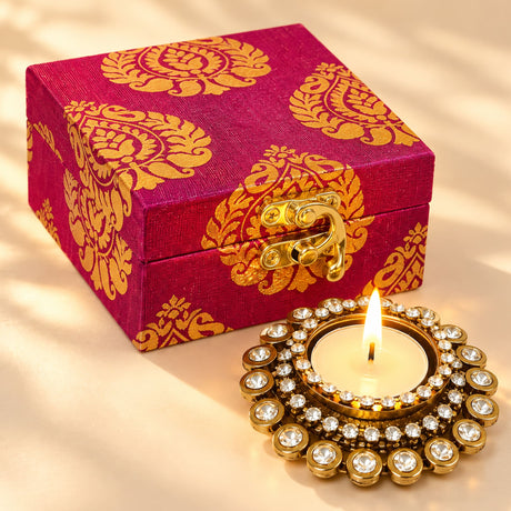A vibrant magenta fabric box with gold paisley patterns and a diamond-encrusted brass diya candle holder for Diwali gift decor