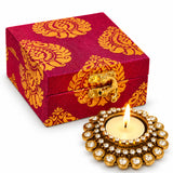 A maroon and gold patterned fabric box with golden clasp beside a diamond-encrusted brass diya candle holder for Diwali gifts