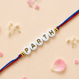 Designer custom named rakhi with white circular beads and gold lettering on red and blue cord for brother and bhabhi