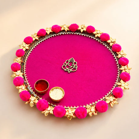 A vibrant pink steel pooja thali with gold floral embellishments bright fuchsia pompoms and central golden ganesha charm