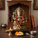 Brass-colored ganesha statue adorned with red and green beaded mala and yellow flowers for pooja and temple decor