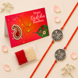 Two ornate silver-toned circular designer floral charm rakhis with orange cords for brother and bhabhi protection gift