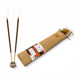 Two dark brown low smoke incense sticks with golden bases burning in a gray ceramic holder next to saffron sandal packaging