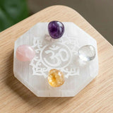A translucent white selenite hexagonal charging plate engraved with an om mandala holding four polished gemstones