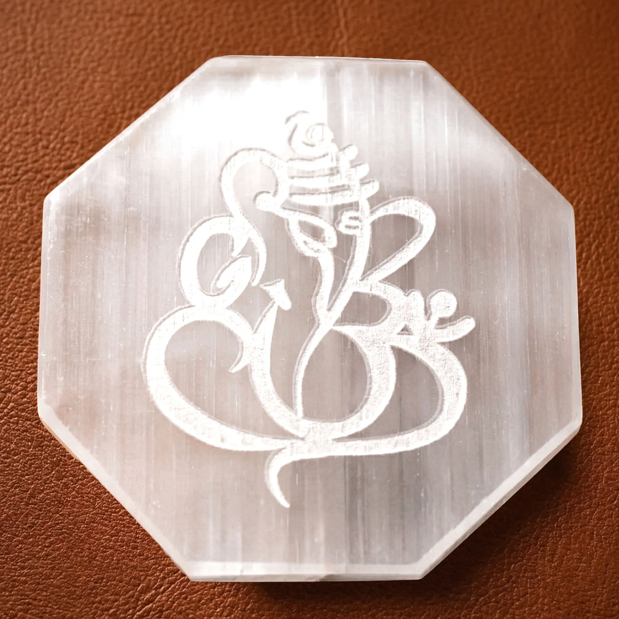 A translucent octagonal white selenite charging plate with a carved ganesha design for crystals and healing stones