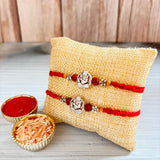 Two red thread designer silver tone ganesha rakhi bracelets with decorative beads displayed on a beige burlap pillow