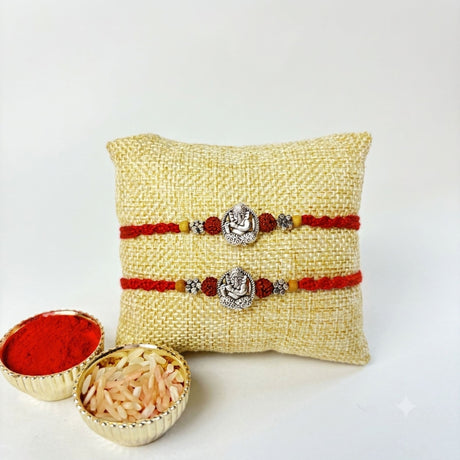 Two red and silver ganesha-themed silver tone rakhi bracelets displayed on a beige woven cushion for brother