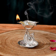 Silver plated peacock diya oil lamp with intricate engravings and lit flame emitting smoke for home pooja mandir