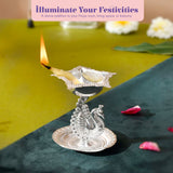 Silver plated peacock diya oil lamp with intricate embossed patterns and lit wick for home pooja mandir and diwali decor