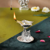 Silver plated peacock diya oil lamp with intricate embossed patterns and lit flame for home pooja mandir and diwali decor