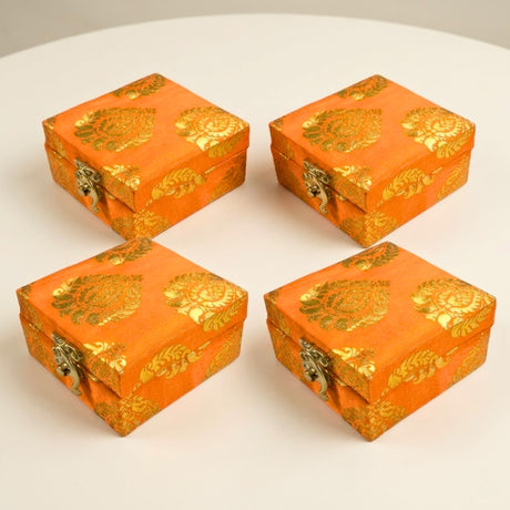 Four vibrant orange fabric boxes with intricate gold gota patti brocade floral patterns and metallic clasps for gifts