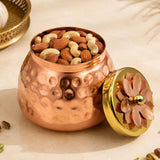 A copper-colored hammered metal nut jar with a golden lid and ornate flower handle for hindu religious gift hamper