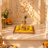 Golden idol of deity seated in yellow attire with intricate jewelry and peacock feather crown on velvet pooja asan pillow set