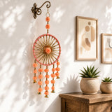 A peach-colored dreamcatcher with a pink frame gold spokes and hanging bells with pom-poms rajasthani wall hanging